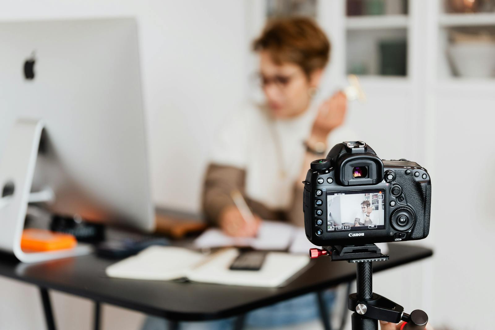 Unrecognizable blurred lady working at table in modern office against photo camera placed on tripod