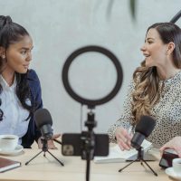 Positive blogger asking questions to African American speaker and recording interview on cellphone at table with microphones